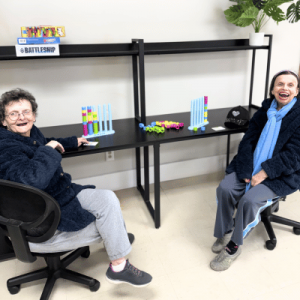 Two seated individuals play a colorful board game on a black table with shelves in a bright room, surrounded by game pieces.