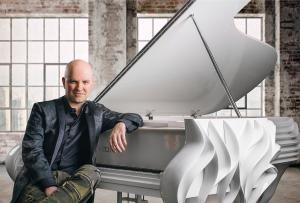 Pianist Martin Mayer sits relaxed beside a white sculptural Fazioli grand piano in a sunlit industrial loft space, wearing a black floral jacquard blazer and olive pants, with exposed brick walls and city views in the background.