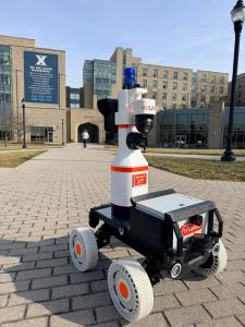 TASKBOT™ SCOUT XT autonomous patrol robot on a campus walkway at Xavier University with buildings in the background.