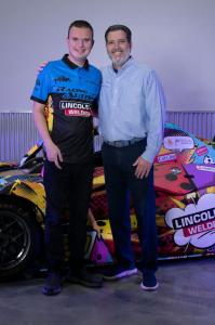Young male racecar driver posing with older man in a light blue buttonup shirt in front of his racecar