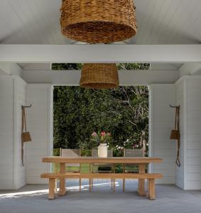 The Savannah Dining Table is accompanied by the Savannah Dining Bench and the Santorini Teak and Mesh Stacking Dining Arm Chairs. They are positioned in the middle back entrance of a pool house, jsut under two large wicker light fixtures. The sun is comin