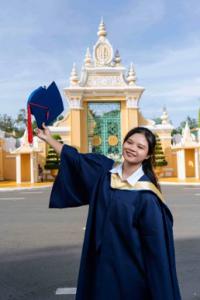 Young Cambodian woman in graduation gown holding cap in front of ornate Phnom Penh architecture after completing university through Love Without Boundaries scholarship program.