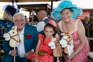 Three guests at a Kentucky Derby themed event smile at the camera while holding plush stick horses. A man in a teal suit with a red rose boutonniere and a woman in a coral floral dress with a large turquoise hat pose beside a young girl in a bright coral