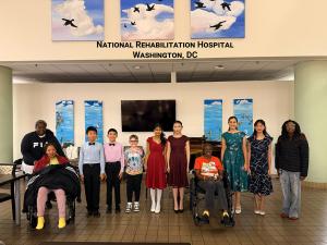 Young EAMA piano students standing with patients in wheelchairs at the National Rehabilitation Hospital in Washington, DC, with colorful bird murals on the wall behind them.