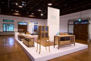 An interior view of a historic exhibition hall with wooden floors and exposed ceiling beams, displaying modern wooden furniture pieces and cabinets on white geometric pedestals for the Daxi Wood Furniture Craft Design Competition.