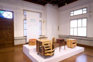 An interior view of a historic exhibition hall with wooden floors and exposed ceiling beams, displaying modern wooden furniture pieces and cabinets on white geometric pedestals for the Daxi Wood Furniture Craft Design Competition.