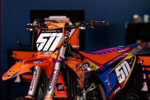 Close-up of an orange KTM motocross bike with number 511 and Dirt Legal decals, photographed indoors against a dark blue background.