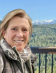 The author photo features a smiling woman, Kristin Whitcomb, standing outdoors on a balcony overlooking a vast forest of evergreen trees with snow-capped mountains in the distance. The sky is clear and bright blue, creating a crisp, refreshing backdrop.