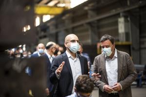 Michael Lassner wearing a face mask while speaking with colleagues inside a large steel manufacturing facility in Spain during the COVID-19 pandemic, with several people in the background also wearing masks.