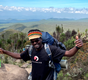 Christopher Sichalwe, a local leader for Follow Alice, smiling and giving a thumbs-up while trekking on Mount Kilimanjaro. He is wearing a black Follow Alice branded t-shirt, a colorful knit beanie, and a professional hiking backpack against a backdrop of
