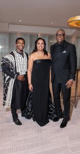 Three individuals pose together at a formal indoor event. The man on the left wears a black traditional-style garment with bold white geometric patterns and black dress shoes. The woman in the center wears a strapless, floor-length black gown with a subtl