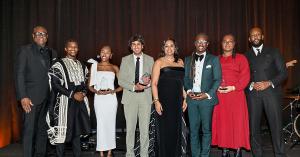Eight honorees stand on stage in formal attire, holding glass awards. Dressed in tuxedos and elegant gowns, they pose before a dark curtain under stage lights, smiling for the camera during the presentation.