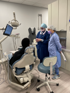 student and instructor stand next to a patient in the dental chair. Both student and instructor are dressed in full PPE gear while the patient sits with their back to the camera.