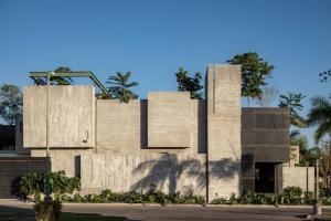 Modern architecture building with exposed concrete facade, cubic volumes, a dark steel entrance element, and surrounding tropical vegetation under clear daylight.