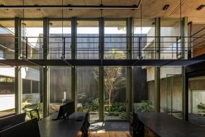 Architecture studio interior with open workstations, a suspended metal walkway, large glass windows, and a central courtyard with plants and natural light.