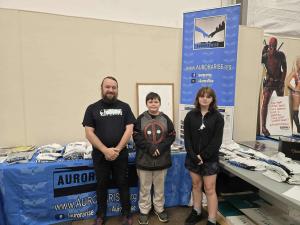 Aurora Rise, a non-profit organization dedicated to supporting victims of the July 20, 2012, theater shooting and fostering community resilience, is proud to announce the opening of the 2026 Aurora Rise Memorial Scholarship application period a man stands in front of a blue tablecloth with 2 scholarship winners