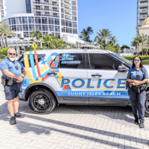 Two police officers stand beside a Sunny Isles Beach police SUV with autism awareness graphics on a sunny day.