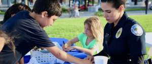 Children interacting with a female police officer at an outdoor event with a blue table and art supplies.