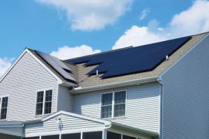As national economic headlines focus on cooling inflation, a divergent reality is taking hold across the Appalachian Highlands A daytime photo of a two-story gray house. A geometric arrangement of dark solar panels covers most of the brown shingle roof, contrasting sharply with the light siding and a bright blue sky filled with puffy clouds.
