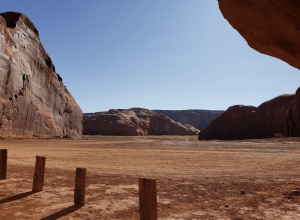 Wide desert landscape in Monument Valley with flat sandy ground, large rock formations, and wooden posts in the foreground.