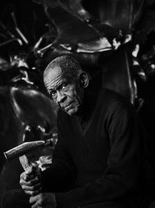 Richard Hunt sitting with a hammer in his hand in front of a bronze sculpture in his studio.