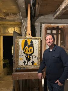 In a moment that underscores Los Angeles’ role as a generator of internationally influential contemporary art, Swiss-American painter and sculptor Sandro Gebert has completed a distinguished residency at the celebrated Château d’Orquevaux Artist & Writers Residency in Orquevaux, France A male artist, smiling, leaning against a painting easel with a contemporary painting of a stylized animal figure in a gold frame.