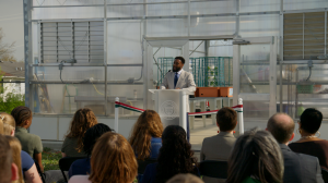 A man speaks from behind a podium in front of a greenhouse.