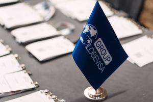 Close-up of a blue Globevisa Group logo flag on a conference table, with professional event badges blurred in the background, representing the global immigration advisory firm.