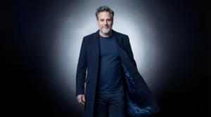 “26 Years on Stage”: A Journey Through His Career A professional portrait of singer and actor Gerónimo Rauch wearing a navy blue coat and t-shirt, looking at the camera against a dramatic studio background with a central light glow.