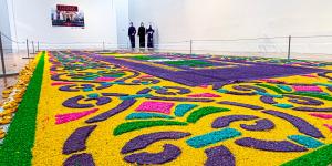 Colorful sawdust carpet installation with intricate patterns displayed across a gallery floor, with visitors standing in the background.