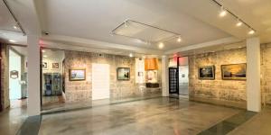 Gallery space with framed landscape paintings displayed on stone-textured walls, with open floor and overhead lighting.