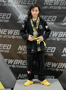 Young athlete Jade Ghezal wearing a black gi with a Winners Academy logo and yellow belt, standing on the podium with a championship medal at a Newbreed Jiu Jitsu Federation competition.