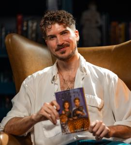 Joey Graceffa poses, seated in a leather library chair, holding a copy of The Subtle Knife by Philip Pullman