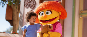A child in a blue dress standing next to a large orange-haired puppet character holding a small brown stuffed animal.