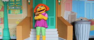 A Muppet character with orange hair and a pink shirt hugs a brown teddy bear while standing on Sesame Street steps.