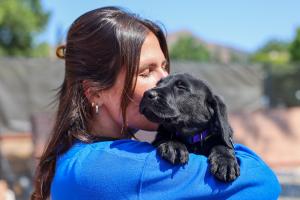 Guide Dogs of America - Puppy Raiser hugging their black labrador puppy for the first time as he soaks up the sunny day with his eyes closes, tilting his head toward the sky.