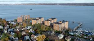 Aerial view of West Shore Towers, a multifamily residential complex of four mid-rise brick buildings situated along the Hudson River waterfront, surrounded by a tree-lined neighborhood with single-family homes, a marina, and distant hills visible across t