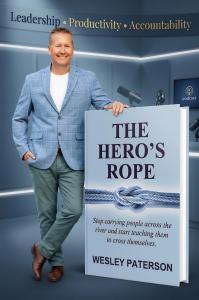Author Wesley Paterson in a blue checked blazer leans beside a large cover of his book “The Hero’s Rope” inside a modern podcast studio, with microphones and a podcast screen in the background under the banner “Leadership - Productivity - Accountability.”