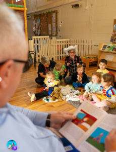 A man reads from a children’s book to a small group of young children seated on the floor in a classroom, with toys, books and a teacher nearby.