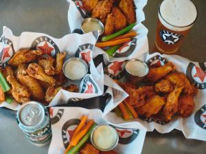 Several baskets of fried chicken wings with dipping sauces, celery, and carrots, served with a draft beer and canned drink on a table at Brake Pad.