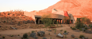 Modern concrete building with unique architecture set in desert landscape with cacti, rocks, and a reddish mountain backdrop at sunset.
