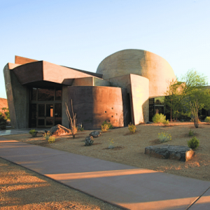 Modern, dome-shaped building with curved concrete walls surrounded by desert landscaping and a clear blue sky.