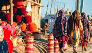 Three women wearing brightly colored shalwar kameez, the photograph shows the back of them, as they are walking past a street stall selling wicker stools