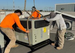 Three technicians from Arrow Appliance Heating & Air Conditioning carefully guide a large commercial AC unit being lowered by a crane onto a rooftop in the Central Valley.