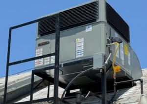 A large grey Trane air conditioning unit mounted on a black metal frame on a residential shingle roof under a clear blue sky.
