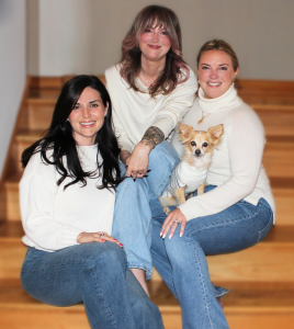 The three co-founders of Meridian Advisory Partners sit together on a wood staircase, all wearing white cream sweaters and jeans. From left: Samantha with dark hair, Amanda in the center with auburn-pink hair, and Melanie on the right with blonde hair pul