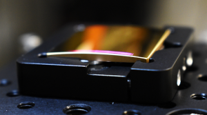 A gold object with pink light coming from a portion of its surface where grating grooves have been ruled into it. The rest of the gold object is shiny and reflective of the light from surrounding objects.