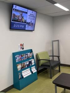 A wall-mounted TV in a clinic waiting room displays a split screen showing a cartoon on the left and a community message with a QR code on the right, alongside a teal brochure rack and seating area.