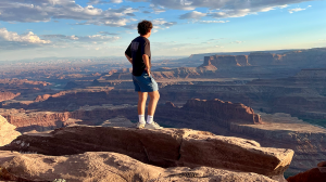 A young person stands on a ledge overlooking a vast Utah canyon landscape under a wide blue sky.