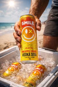 Hand holding Smile Spirits Pineapple Orange Vodka Soda can over ice at a sunny beach scene